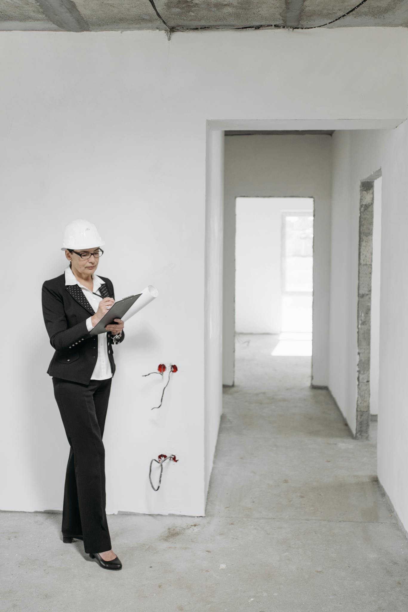 Female architect in hard hat inspecting an unfinished building interior with formal attire and clipboard.